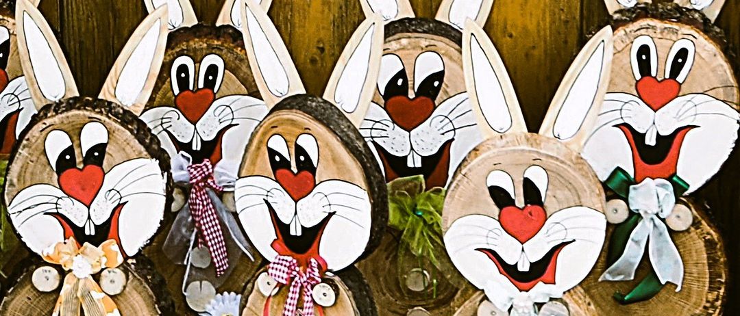Eine Sammlung geschnitzter Holzhasenfiguren mit großen Ohren, gemalten Gesichtern und dekorativen Bändern. Jeder Hase hat eine einzigartige Miene und Accessoires wie Knöpfe und Blumen. Sie sind auf einem hölzernen Hintergrund platziert.