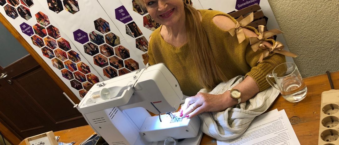 A woman with blonde hair is smiling while sewing on a sewing machine. She has a wristwatch and ribbons on her shoulders. She is sitting on a wooden desk with a glass of water, a piece of paper, and a box. Behind her is a wall with a collage of photos.