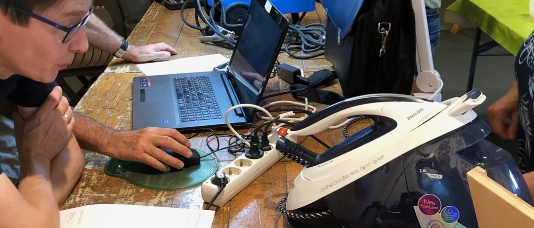 Mehrere Personen arbeiten an einem Tisch mit Laptops und elektronischen Geräten. Eine Person mit Brille konzentriert sich auf den Laptop-Bildschirm.