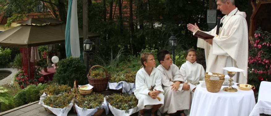 Ein Priester führt eine Zeremonie mit drei Kindern durch, die neben Körben mit Pflanzen sitzen, unter einem überdachten Bereich mit Bäumen und einer Flagge im Hintergrund.
