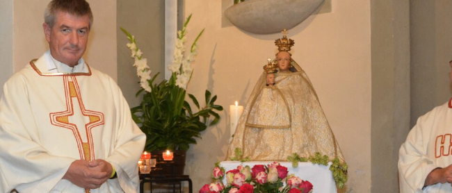 Zwei Priester stehen in der Nähe eines mit Blumen und Kerzen geschmückten Altars, hinter ihnen eine Statue einer Heiligen mit Krone.