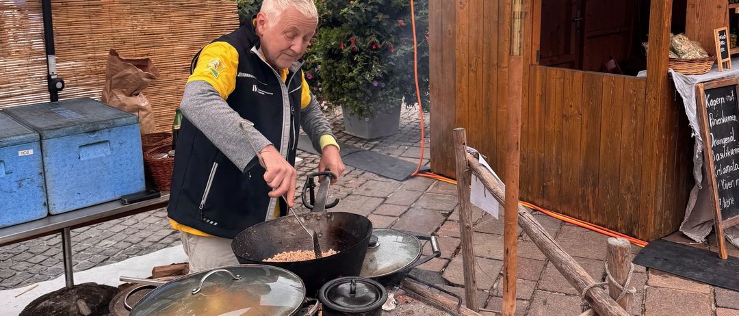 Ein älterer Mann kocht auf einem Feuerplatz vor einem Imbissstand. Der Stand hat Schilder für 'Gemüse-Pfanne' und 'Dinkel-Grief-Heiden-Sterz'. In der Nähe sind Lagerboxen und Körbe.