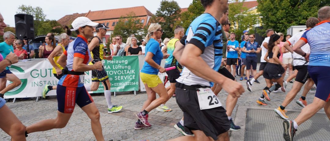 Läufer nehmen an einem Marathon auf einer Kopfsteinpflasterstraße teil, während Zuschauer zuschauen. Ein Banner steht hinter ihnen, und Gebäude mit Bäumen sind im Hintergrund.