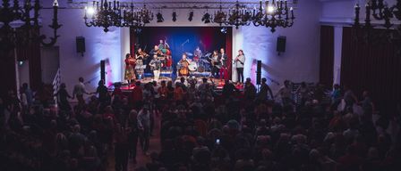 A lively music performance on stage with a band of violinists, cellists, and a saxophonist. The audience is seated in front, some standing and dancing. The stage is illuminated with blue and red lights, and chandeliers are hanging from the ceiling.
