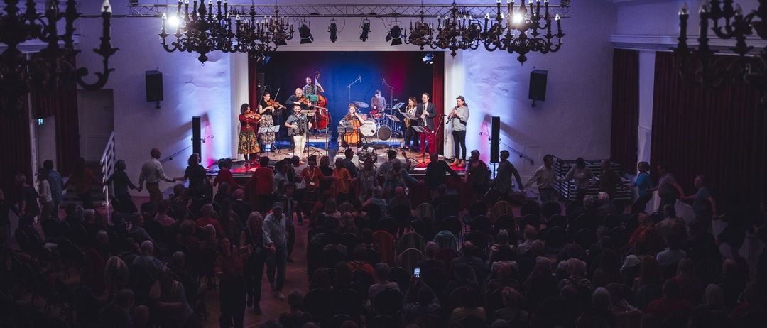 Ein lebhaftes Musikkonzert auf der Bühne mit einer Band aus Geigern, Cellisten und einem Saxophonisten. Das Publikum sitzt vorne, einige stehen und tanzen. Die Bühne ist mit blauen und roten Lichtern beleuchtet, und Kronleuchter hängen von der Decke.