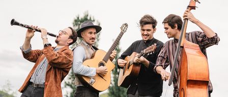 Four men stand on a grassy field, smiling, and playing musical instruments. One man plays the clarinet, another plays a guitar, and two others play a cello and another guitar. Behind them are trees and buildings.