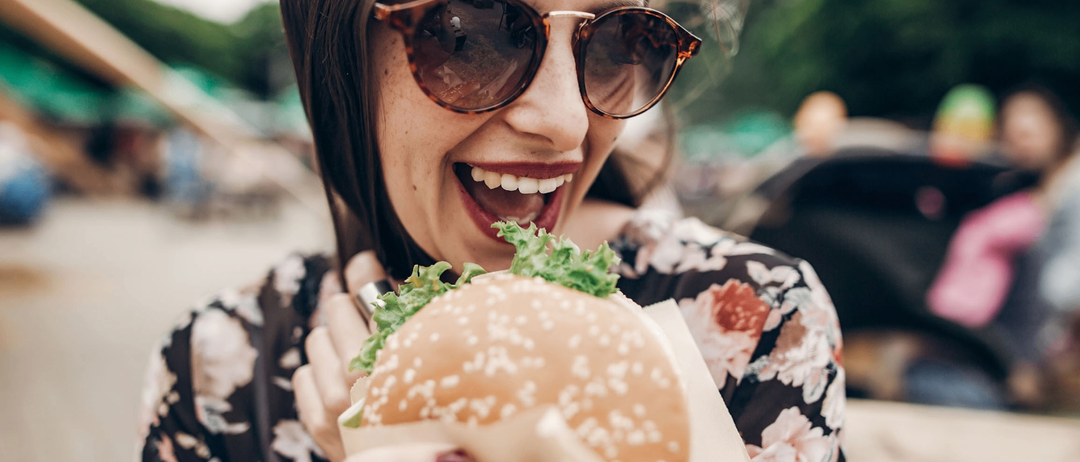 A woman with sunglasses is holding a hamburger, smiling, and appears to be enjoying it. She is wearing a floral dress.