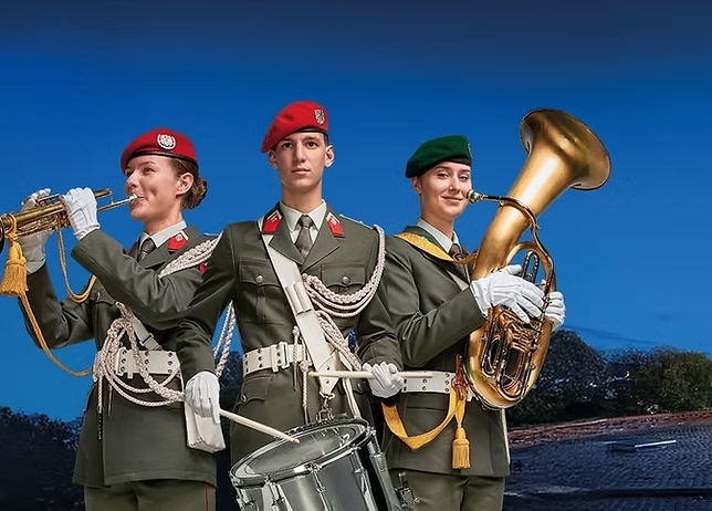 Drei Mitglieder einer Militärkapelle in grünen Uniformen und Mützen halten Trompete, Trommel und Tuba und stehen vor einem blauen Himmel.