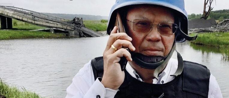 Ein Mann in einem Presse-Weste und Helm mit TV darauf hat ein Telefongespräch in einem Wassergebiet.