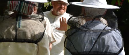 Eine Gruppe von Imkern in Schutzkleidung steht im Freien, einer gestikuliert, während die anderen zuschauen.