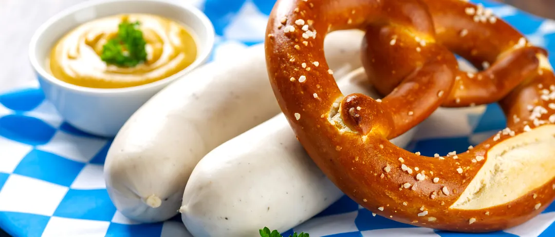 Ein Brezel mit Sesamkörnern und zwei Würsten auf einem blau-weiß karierten Tuch. Ein Senfschälchen ist ebenfalls vorhanden.