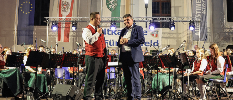 Two men stand on stage in front of an orchestra, holding microphones. Behind them, a large banner is displayed. The audience sits on chairs and watches.