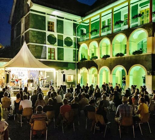 Eine musikalische Aufführung findet im Freien vor einem Gebäude mit grünen Lichtern statt. Das Publikum sitzt auf Stühlen und beobachtet die Musiker. Das Zelt befindet sich auf der linken Seite der Bühne.
