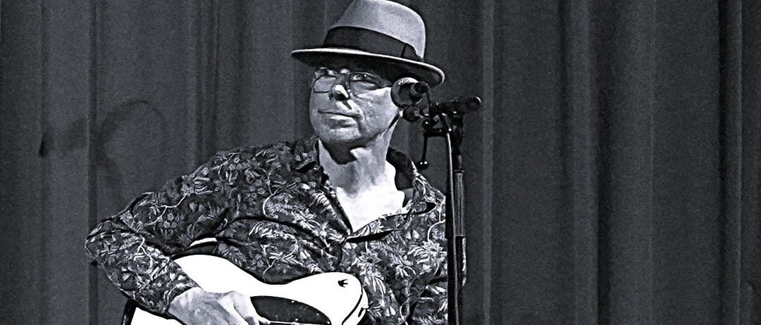 Ein Mann mit Hut und Brille spielt eine weiße Gitarre auf der Bühne. Er sitzt auf einem Hocker mit einem Mikrofon vor ihm.