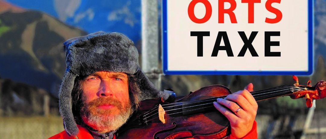 Ein Mann in einer roten Jacke und einem Pelzhut spielt eine Geige vor einem Schild, das 'Orts Taxe' steht. Hinter ihm sind Berge unter einem klaren blauen Himmel sichtbar.
