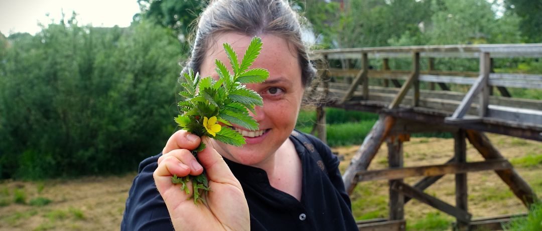 Eine Frau hält eine gelbe Blume in der Hand, lächelt, mit einer Holzbrücke und Grünpflanzen im Hintergrund.