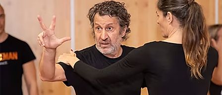 A man and a woman are dancing together, the man is making a hand gesture while the woman is looking at him. They are both wearing black clothes.