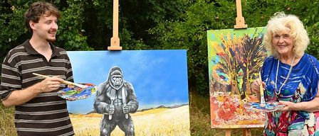 Ein Mann und eine ältere Frau malen im Freien auf Staffeleien. Der Mann malt einen Gorilla und die Frau ein farbenfrohes abstraktes Bild. Das Bild hat einen Wasserzeichen mit dem Text 'Farben der Generationen'.