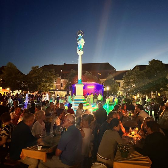 Eine nächtliche Zusammenkunft von Menschen um Tische in einem Außenbereich, mit einer von bunten Lichtern beleuchteten Statue im Hintergrund.