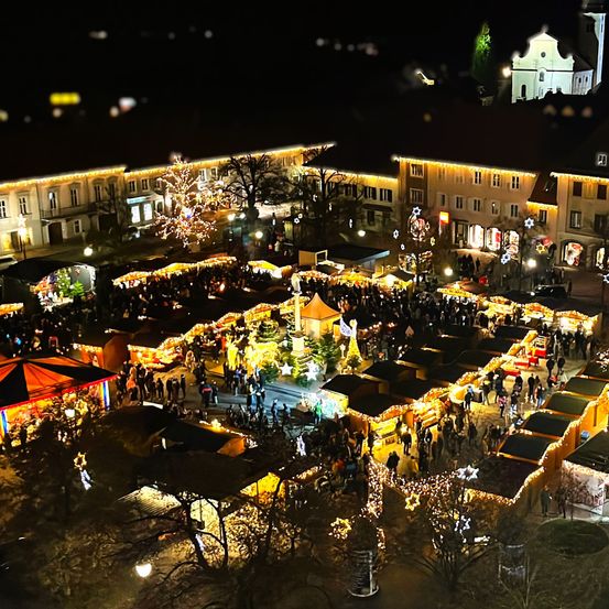 Ein nächtlicher Luftblick auf einen festlichen Weihnachtsmarkt mit zahlreichen Ständen, einem geschmückten Baum und einer Menschenmenge. Im Hintergrund sind Gebäude mit Lichtern geschmückt.