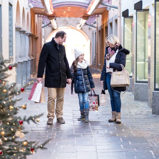 Eine dreiköpfige Familie, ein Mann, eine Frau und ein junges Mädchen, gehen mit Einkaufstüten einen gepflasterten Weg entlang, in der Nähe eines Weihnachtsbaums und Geschäften.