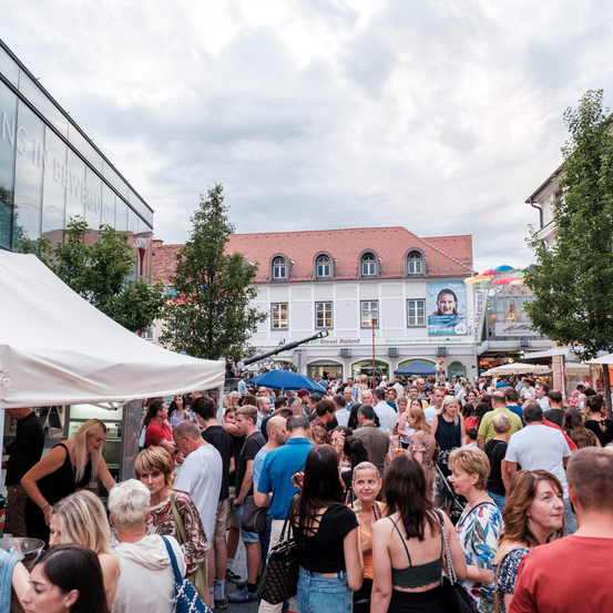 Eine Menschenmenge versammelt sich auf einem Außengelände, wahrscheinlich bei einem Festival. Es gibt Zelte, ein Gebäude mit rotem Dach und ein großes Plakat. Der Himmel ist bewölkt.
