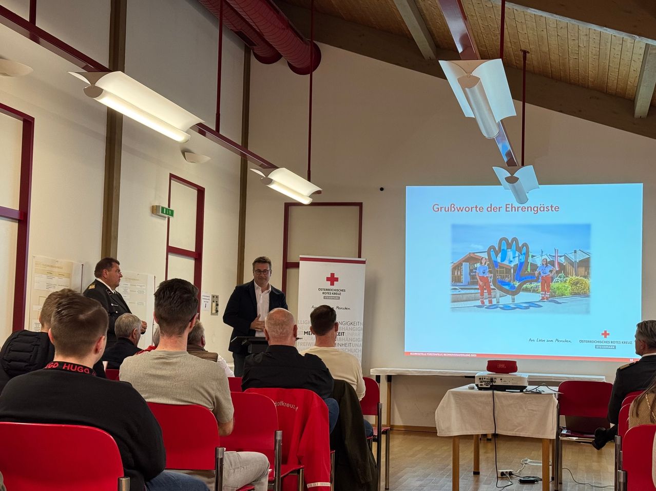 Ein Mann spricht an einem Rednerpult vor einer Gruppe von Menschen, die in roten Stühlern sitzen. Eine Leinwand hinter ihm zeigt eine Grafik eines Gebäudes.