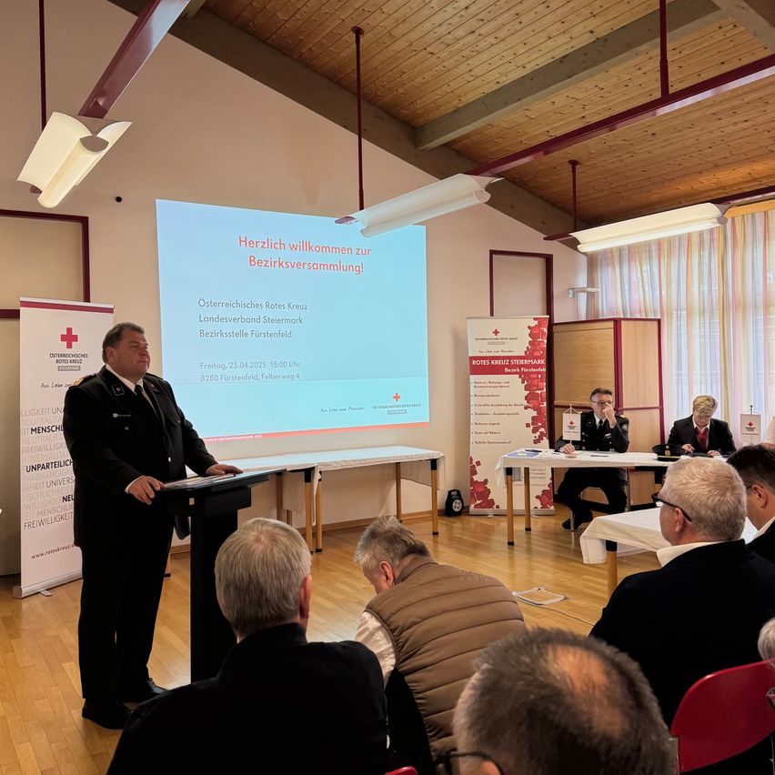 Ein Mann hält eine Rede bei einer Versammlung in einem Konferenzraum mit einer Projektionsleinwand. Die Leute sitzen vor ihm.