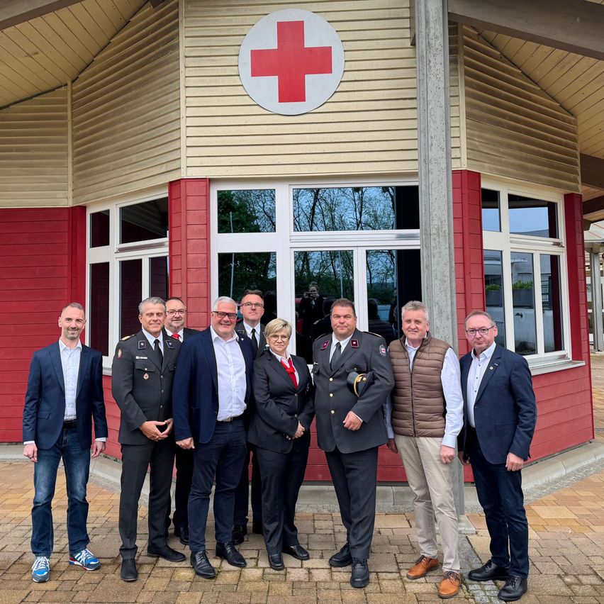 Eine Gruppe von acht Männern und Frauen, gekleidet in formeller Kleidung, steht vor einem roten Gebäude mit einem weißen Kreuz. Das Gebäude hat Glasfenster und ein weißes Kreuz an seiner Wand.