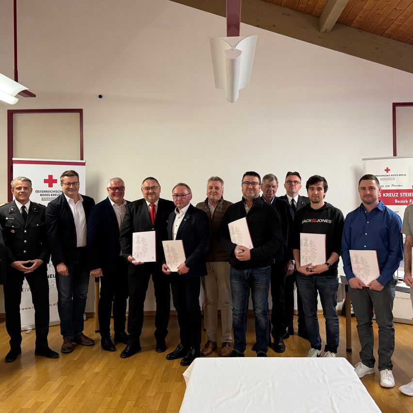 Elf Männer stehen auf einem Holzboden und halten Urkunden in den Händen. Hinter ihnen ist ein weißer Stoff auf einem Tisch ausgebreitet. Dahinter ist eine Fahne an der Wand befestigt.