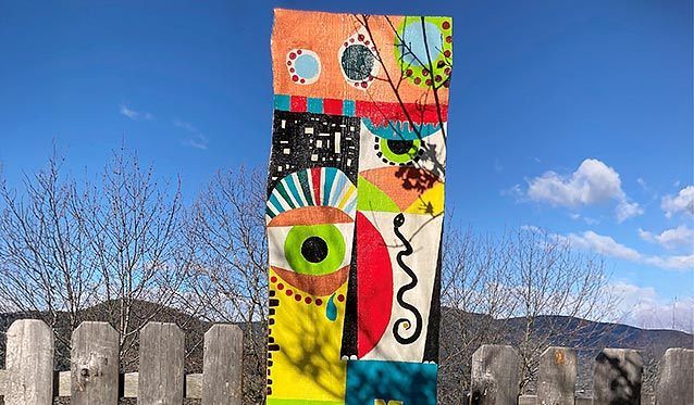 Ein farbenfrohes abstraktes Gemälde steht auf einem Holzzaun vor einem Hintergrund aus klarem blauem Himmel mit flauschigen Wolken und einer entfernten Bergkette.