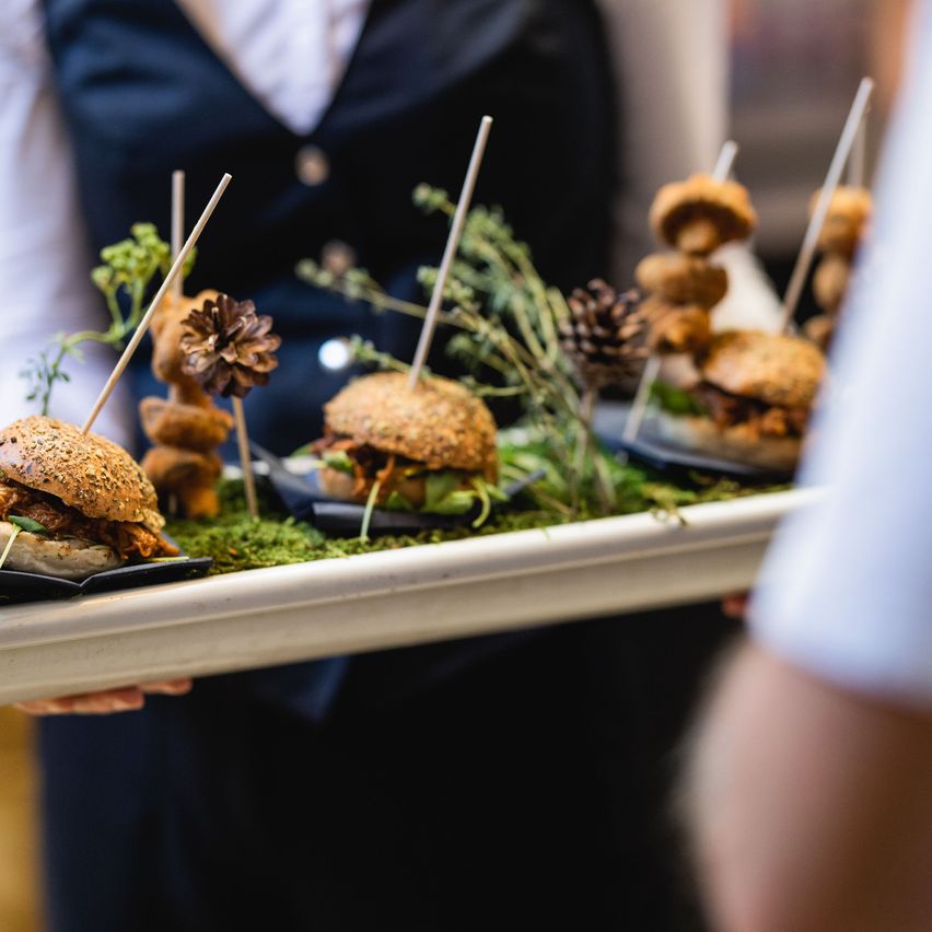 Ein Kellner hält eine Tablett mit Vorspeisen mit Burger-Slidern und Hähnchenspießen mit Kiefernzapfen.