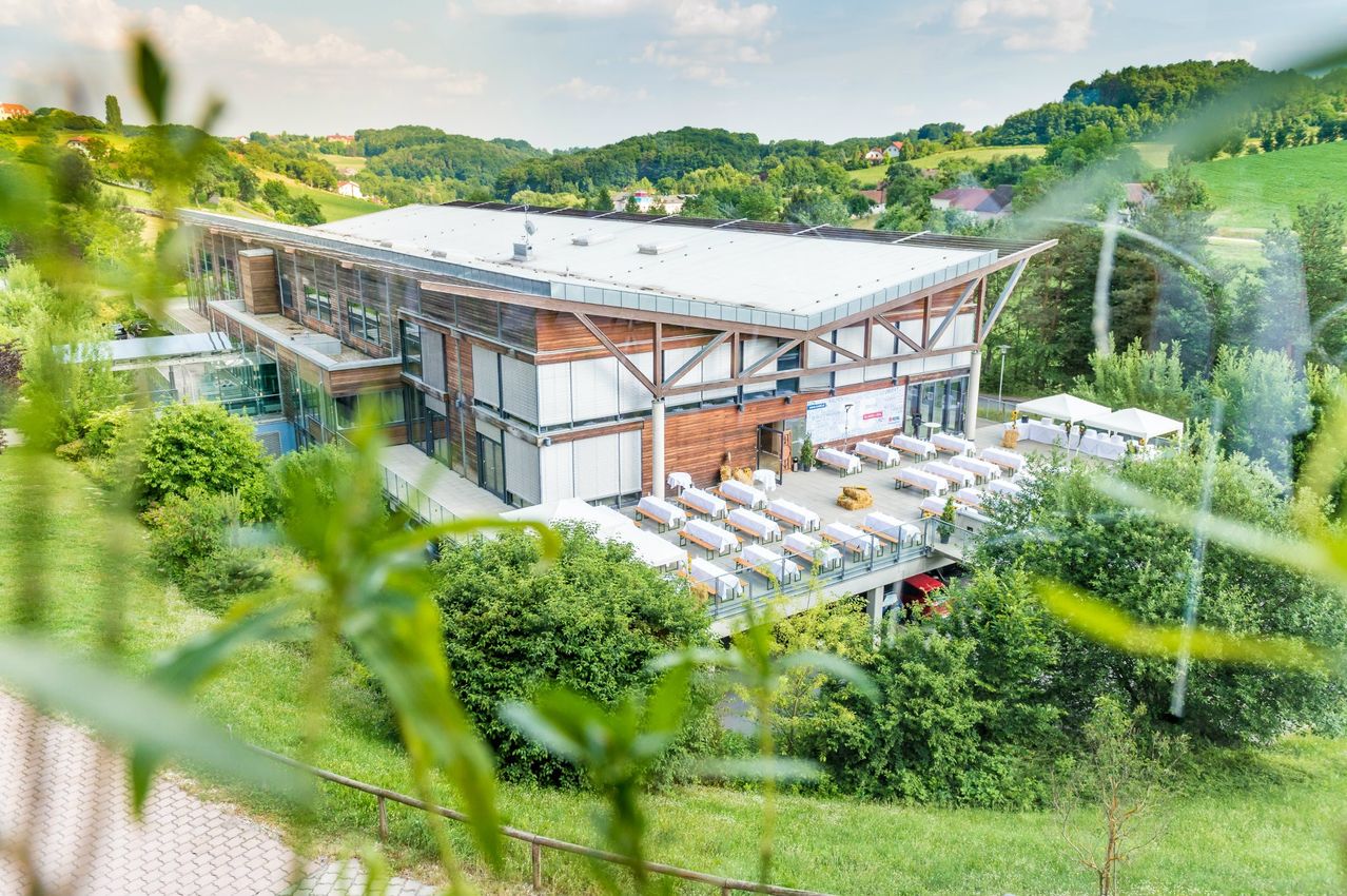 Ein Blick von oben auf ein großes Gebäude mit flachem Dach und mehreren weißen Tischen und Stühlen auf der Terrasse. Das Gebäude ist von üppiger Grünfläche und Bäumen umgeben.