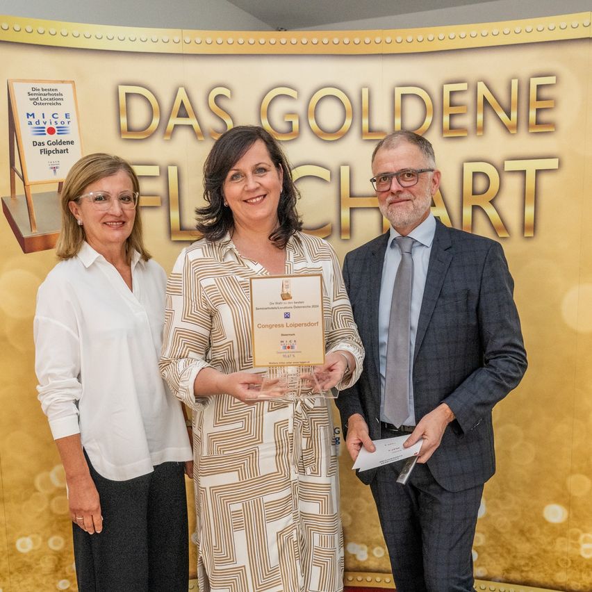 Drei Personen stehen vor einem Banner mit goldenen Buchstaben, auf dem Das Goldene Flipchart steht. Die Frau in der Mitte hält eine Urkunde und lächelt, während der Mann rechts ein Papier in der Hand hält und darauf schaut.