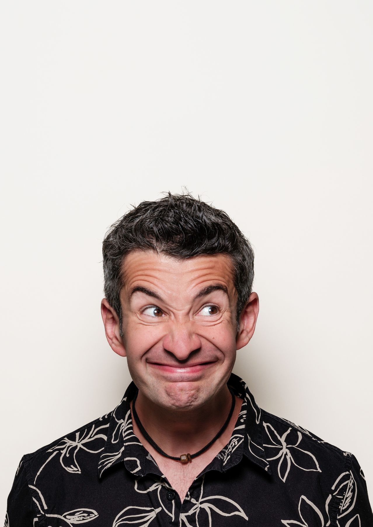 Nahaufnahme des Gesichts eines Mannes mit kurzen Haaren, schwarzer Blumenbluse und Halskette. Er macht eine lustige Grimasse.