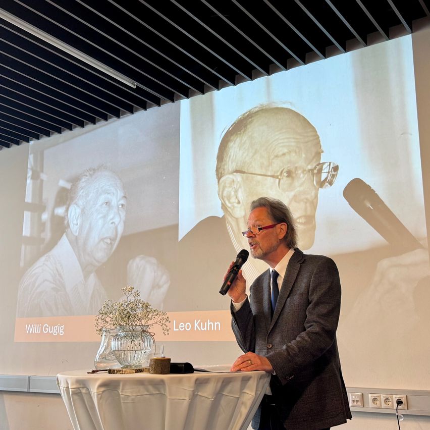 Ein Mann in einem Anzug spricht in ein Mikrofon. Ein Projektionsbild hinter ihm zeigt zwei Fotos, eines von Leo Kuhn und eines von Willi Gugig.