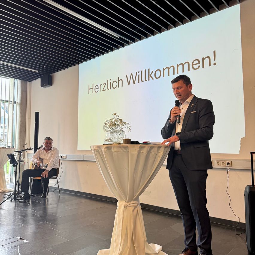 Ein Mann steht hinter einem Tisch und spricht in ein Mikrofon. Ein anderer Mann sitzt in der Nähe und spielt Gitarre. Eine große Leinwand hinter ihnen zeigt die Worte 'Herzlich Willkommen!'.