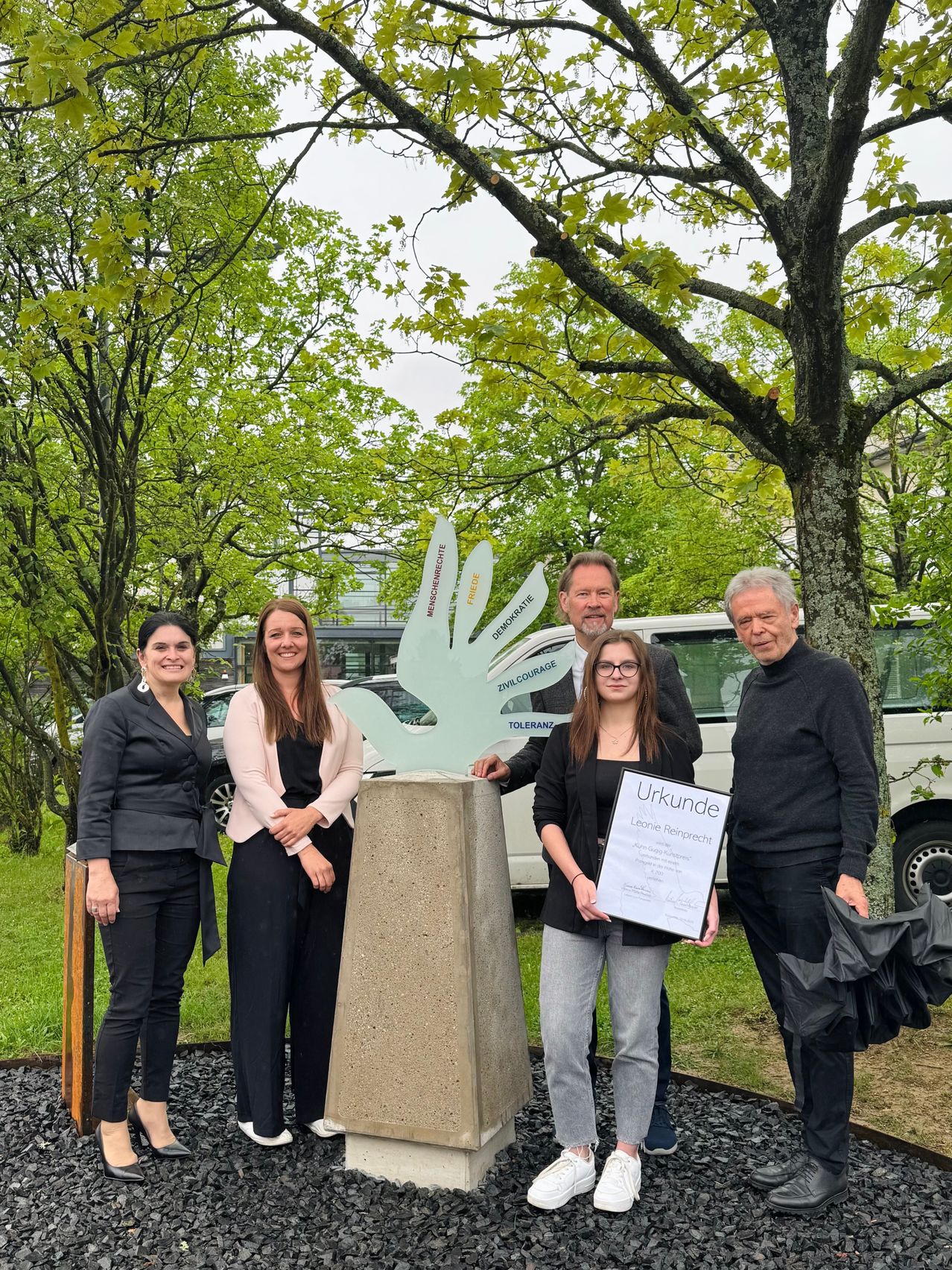 Fünf Personen stehen vor einer Skulptur. Eine Frau hält eine Plakette mit der Aufschrift 'Urkunde'. Im Hintergrund sind grüne Bäume und ein weißer Van zu sehen.