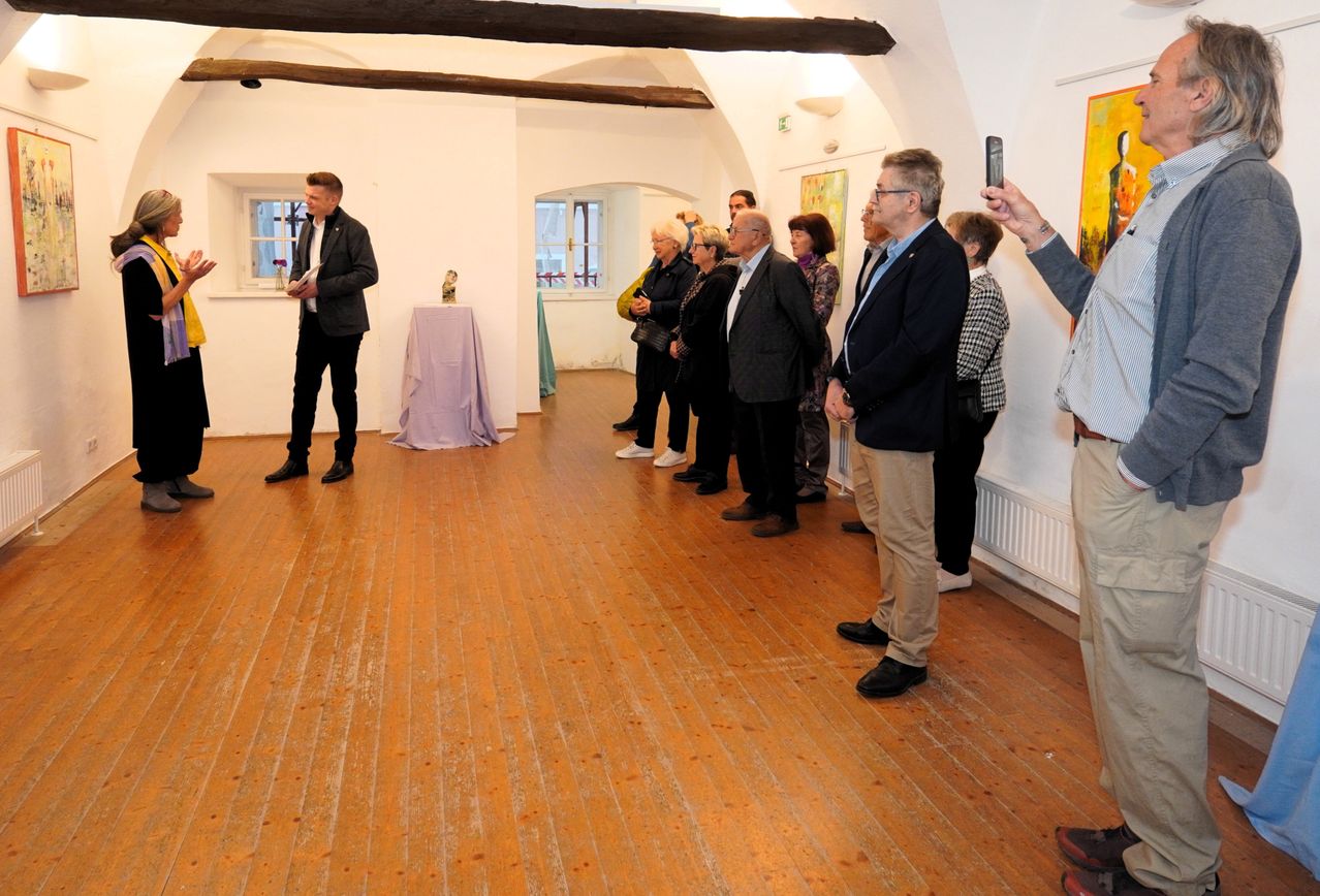 Ein Mann spricht zu einer Gruppe in einer Kunstgalerie. Andere hören zu, einige machen Fotos. Der Raum hat Holzböden und weiße Wände.