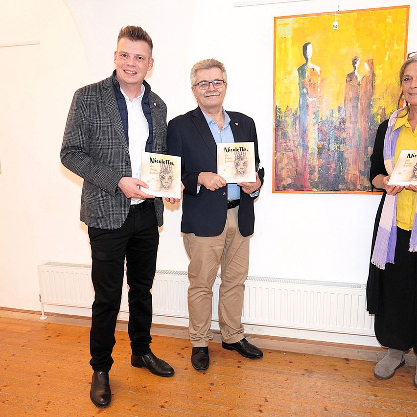 Drei Menschen stehen zusammen, halten Bücher und einer trägt eine Brille, ein Gemälde ist an der Wand.