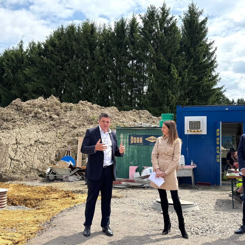 Ein Mann und eine Frau stehen vor einer Baustelle. Der Mann spricht und gestikuliert mit den Händen. Dahinter befindet sich ein blaues Gebäude und ein Haufen Erde.