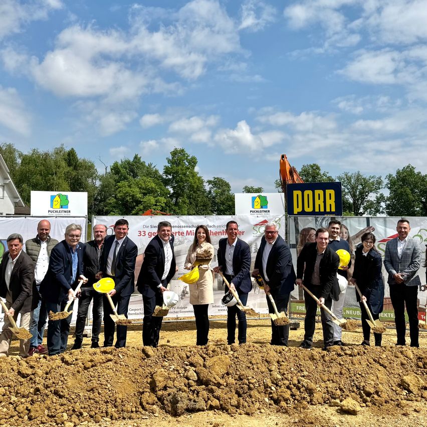 Eine Gruppe von Männern und Frauen posiert für ein Foto, während sie Schaufeln halten, bei einer Grundsteinlegungszeremonie. Hinter ihnen ist ein großes Banner mit dem Wort 'PORR' zu sehen. Die Veranstaltung findet im Freien statt, mit einem Haufen Erde und einem sonnigen Himmel im Hintergrund.