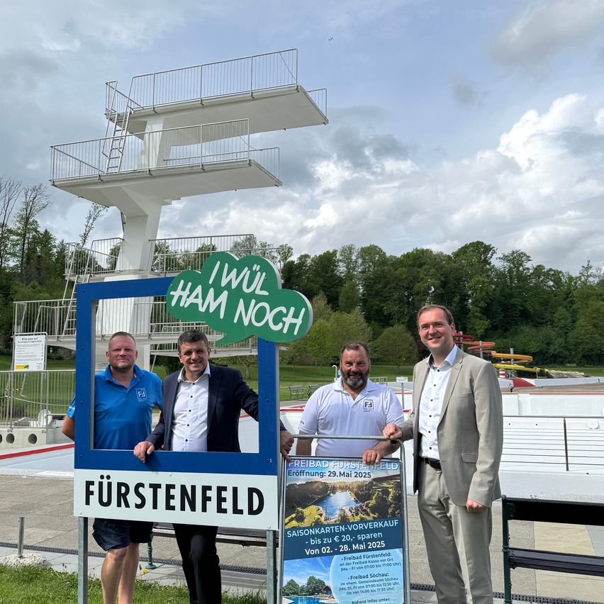 Vier Männer posieren lächelnd vor einem Pool. Ein Schild mit der Aufschrift 'Fürstenfeld' und einer Wolke darüber. Dahinter ist ein Sprungturm und üppige Grünflächen zu sehen.
