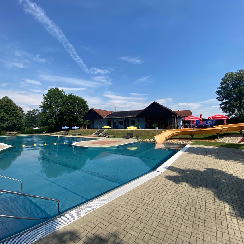 Ein sonniger Tag an einem Freibad mit Wasserrutsche, Liegestühlen und Sonnenschirmen. Der Pool ist von Bäumen und einem Gebäude im Hintergrund umgeben.