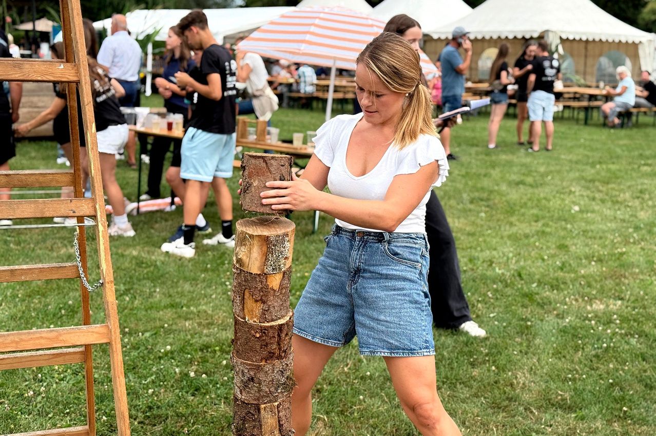 Eine Frau in Jeansshorts und einem weißen Oberteil hackt Holz bei einer Outdoor-Veranstaltung. Hinter ihr stehen und sitzen Leute unter Zelten. Tische mit Getränken und Essen sind aufgebaut.