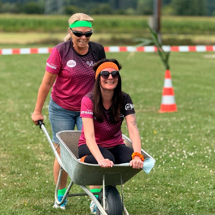 Zwei Frauen in Sportkleidung, eine schiebt einen Schubkarren, in dem eine andere sitzt, auf einer Wiese mit einem Verkehrskegel und einem unscharfen Hintergrund.
