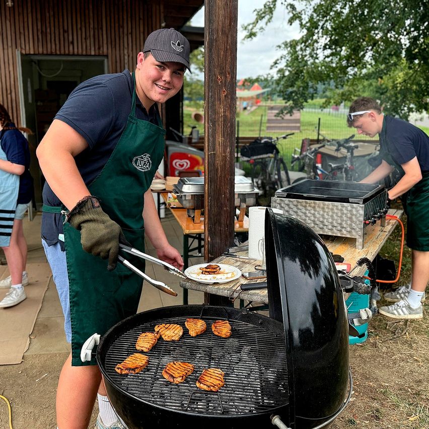 Ein Mann kocht im Freien auf einem Grill, trägt eine Schürze und Handschuhe. Hinter ihm kocht eine andere Person auf einem separaten Grill. Fahrräder sind in der Nähe geparkt, und ein Gebäude ist im Hintergrund sichtbar.