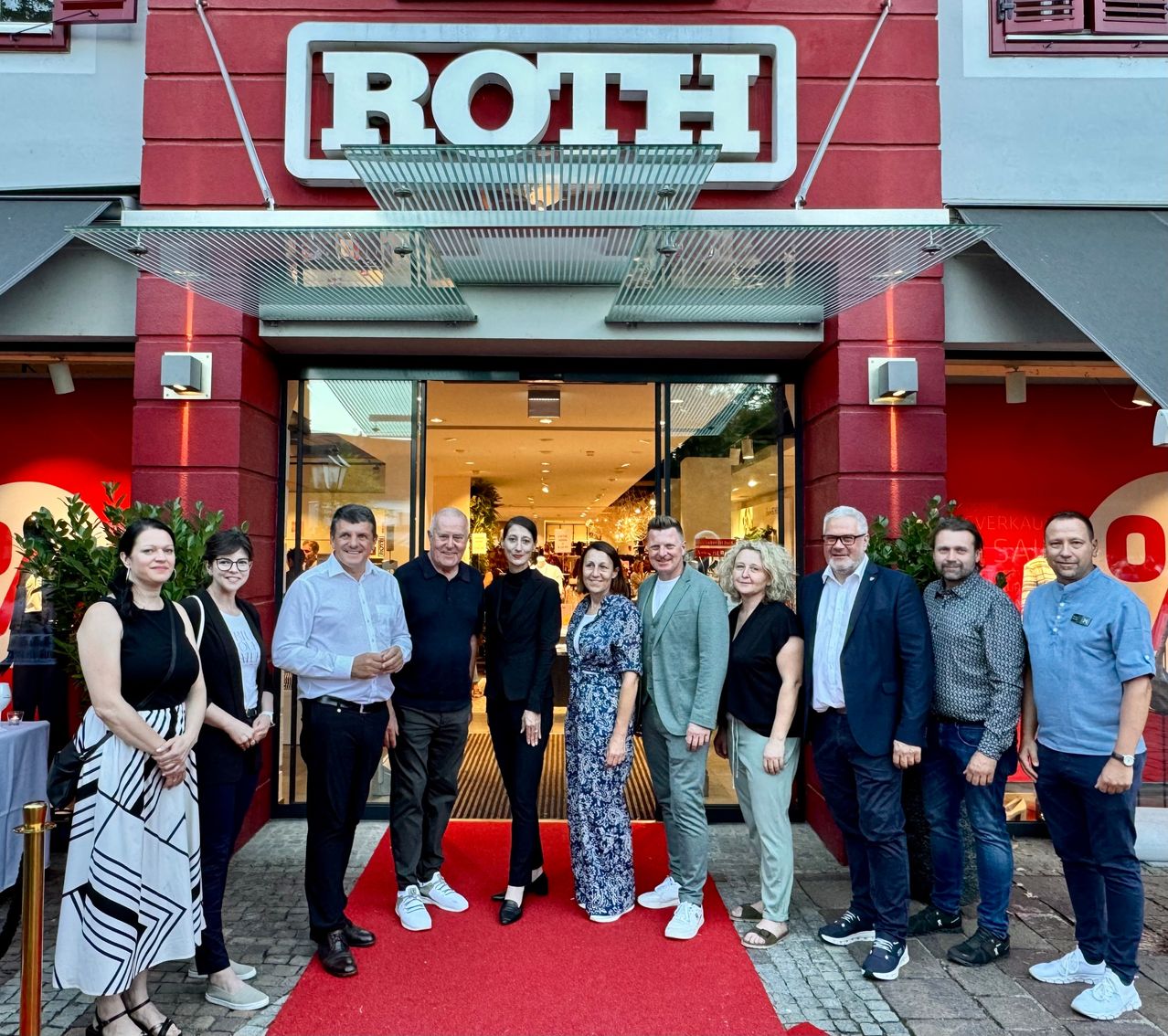 Eine Gruppe von Menschen steht vor einem Geschäft mit einem roten Teppich und einem roten Schild mit der Aufschrift ROTH.