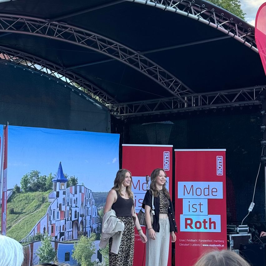Zwei Frauen stehen auf einer Bühne vor einer großen Kulisse mit einem farbenfrohen Schloss und Berglandschaft. Sie tragen Sommerkleidung und scheinen zu lächeln und für ein Foto zu posieren.