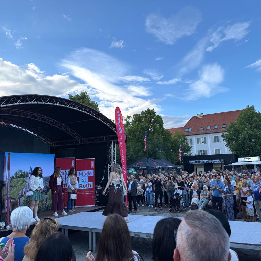 Eine Gruppe von Menschen auf der Bühne, mit drei Personen vorne, möglicherweise bei einer Aufführung oder Rede. Eine große Menschenmenge versammelt sich um sie herum, einige halten Kameras in der Hand. Der Hintergrund zeigt ein großes Banner mit der Aufschrift 'Mode ist Roth'.